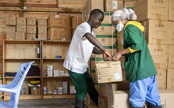 Deux employés de MSF déplacent des cartons contenant des trousses contre le choléra dans un local où sont stockés de nombreux autres cartons empilés contre un mur.