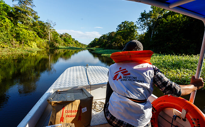 Un membre du personnel de MSF portant un gilet de sauvetage est assis à l’avant du bateau qui descend une rivière traversant la forêt.