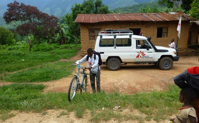 Une personne marche avec un vélo devant une maison en adobe devant laquelle est garé un camion de MSF.